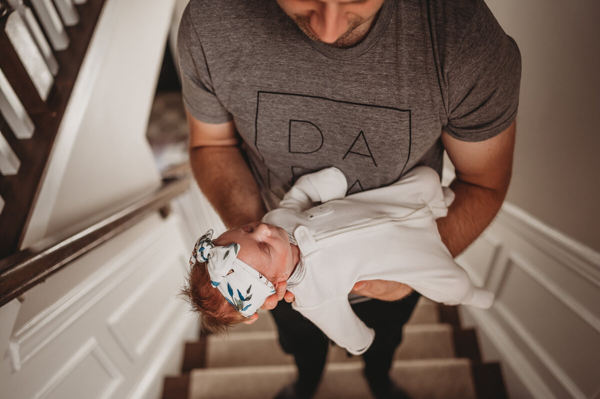 Dad holding newborn on stairs in their house wearing a dad shirt