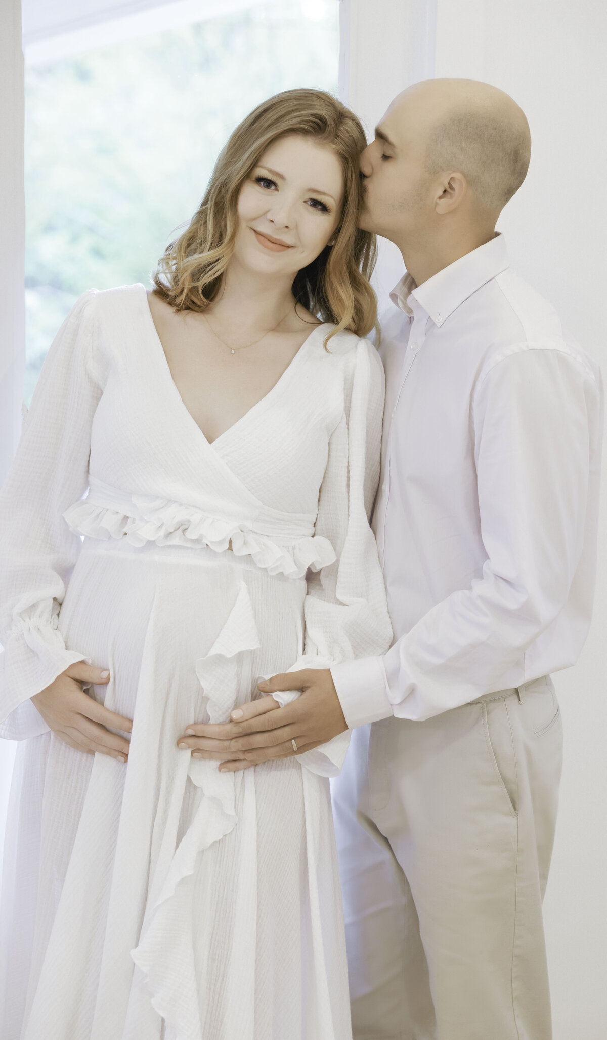 Pregnant woman in white dress posing with husband in studio