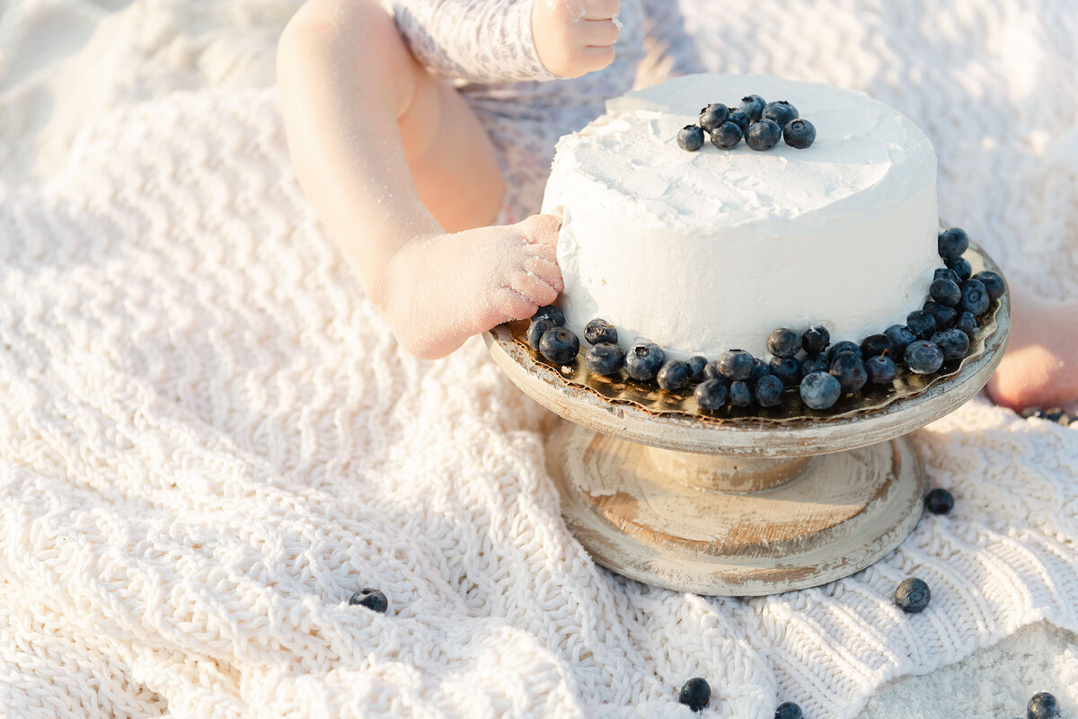 A baby on the beach sticking her toe into her cake smash with blueberries.