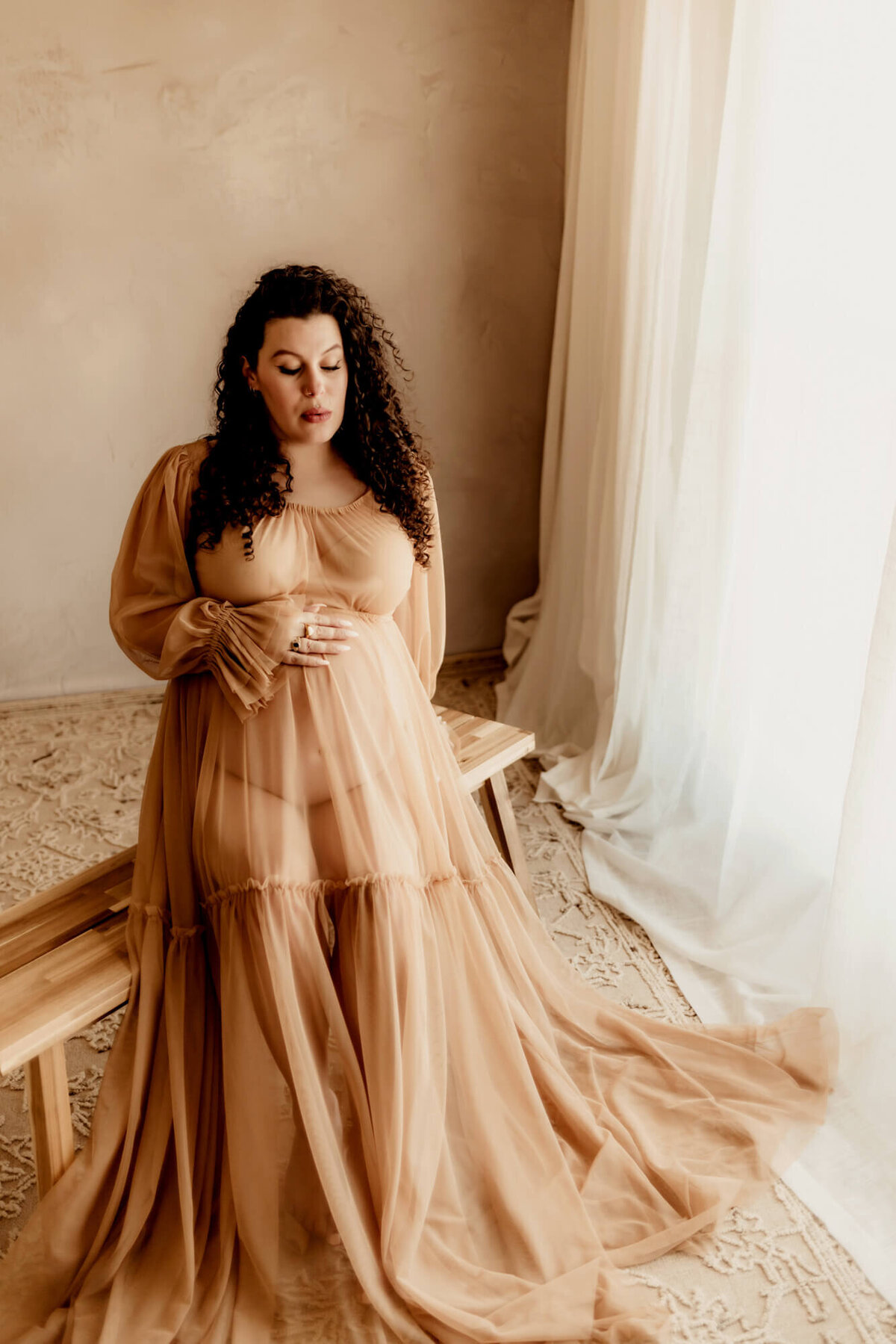Pregnant mother with curly hair wearing a sheer tan dress sitting on a bench and looking at her baby bump.