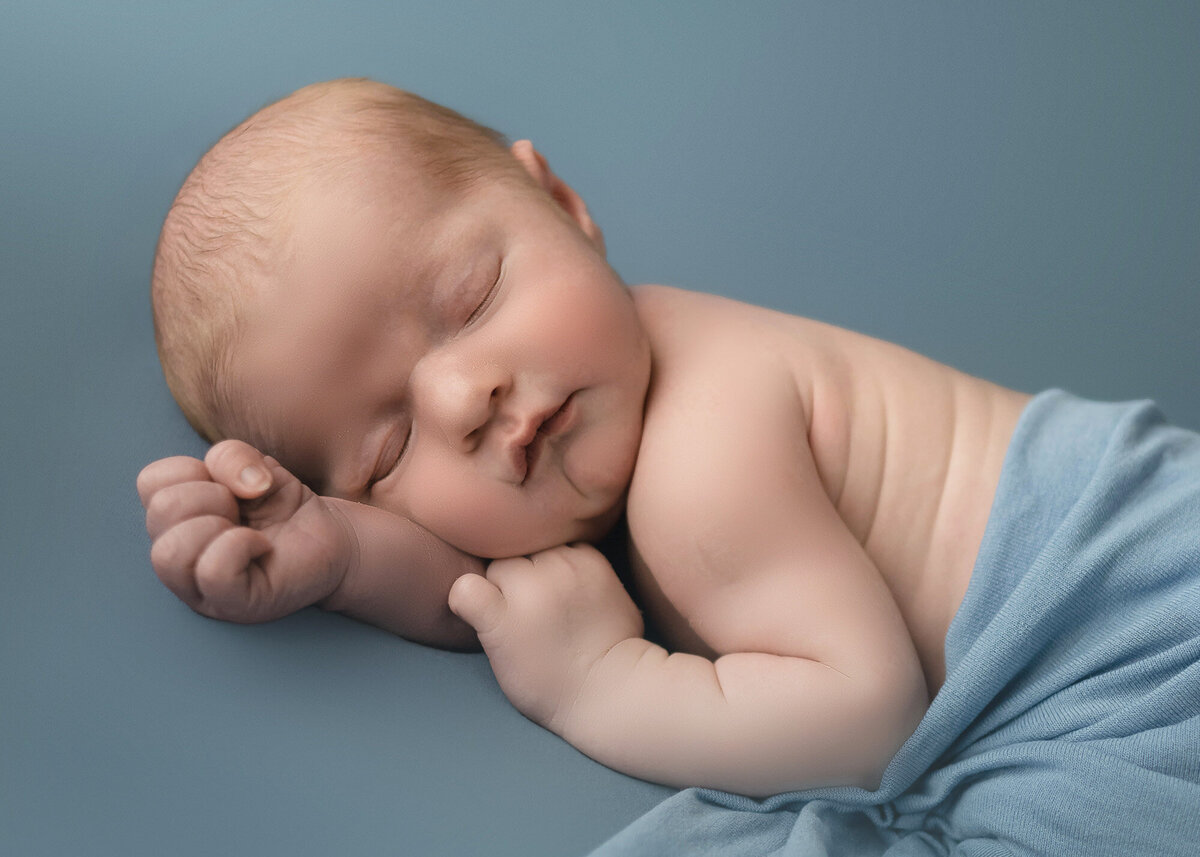 A newborn baby boy sleeps with fist clenched under a blue blanket in the studio of a Pella Newborn Photographer