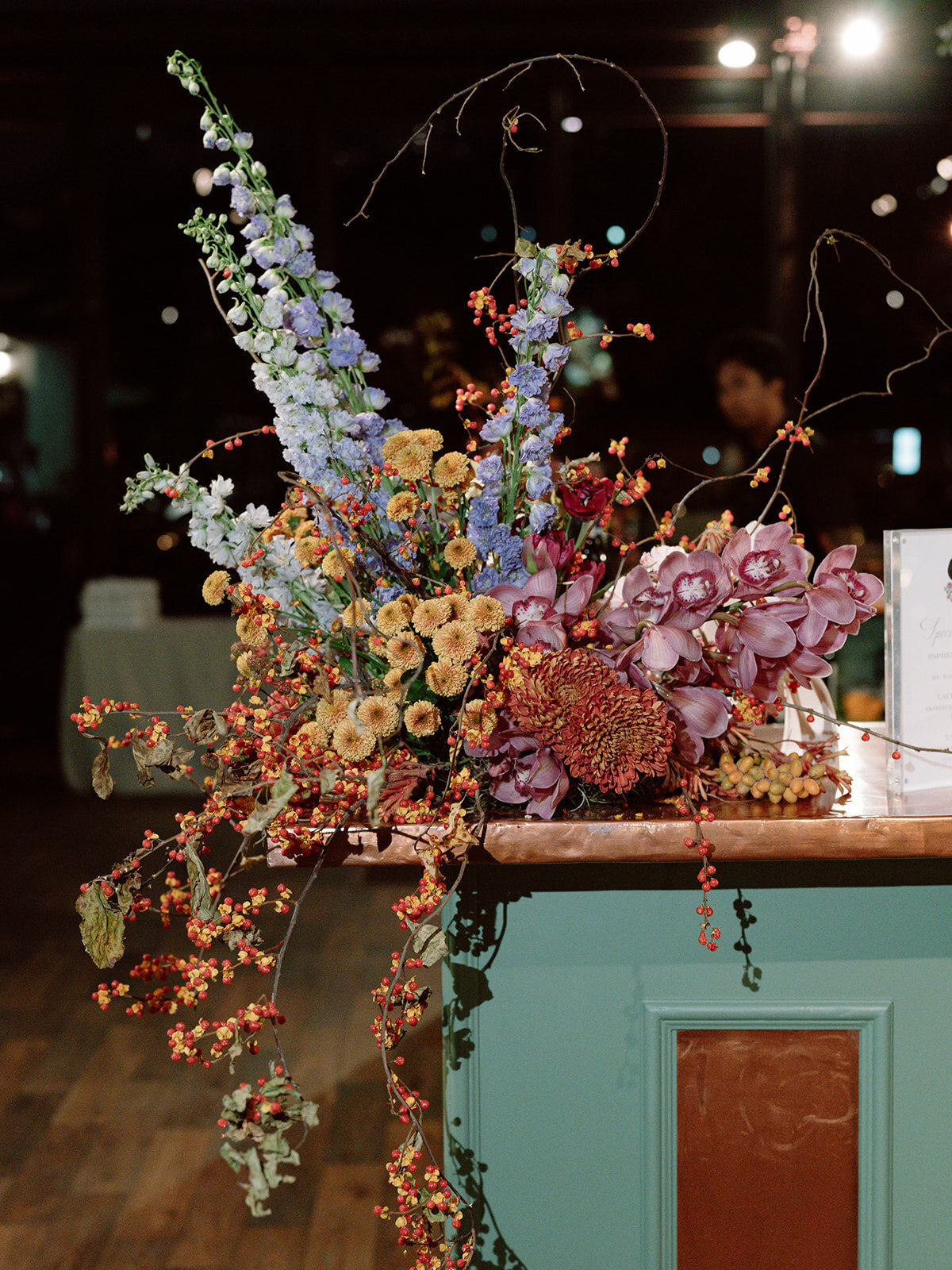 Trailing bar meadows with florals in hues of purple, ruby, copper, gold, and rust for fall wedding reception. Sculptural product clustered floral design for fall wedding. Product clustered floral statements in a tasteful fall color palette of copper, ruby, gold, burgundy, rust, and lavender. Orchids, bittersweet berries, butterfly ranunculus, and tulips are highlighted. Unique floral design with flower forward large scale installations. Design by Rosemary & Finch Floral Design in Nashville, TN.