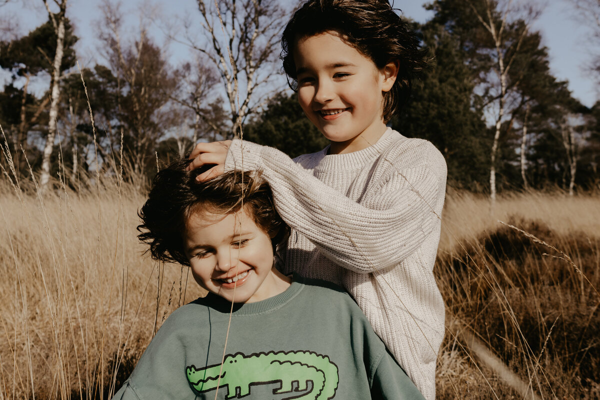 Twee kinderen staan in een veld, omringd door gras en bloemen, met een heldere lucht op de achtergrond.