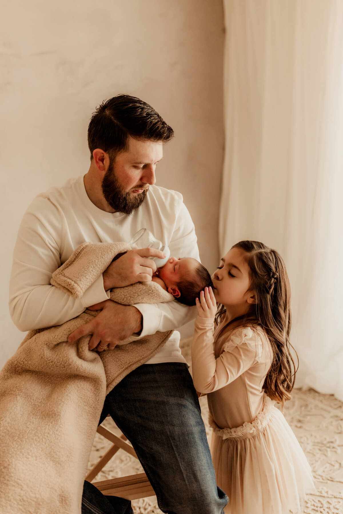 Father bottle feeds his baby boy while his older daughter kisses her little brother on the forehead.