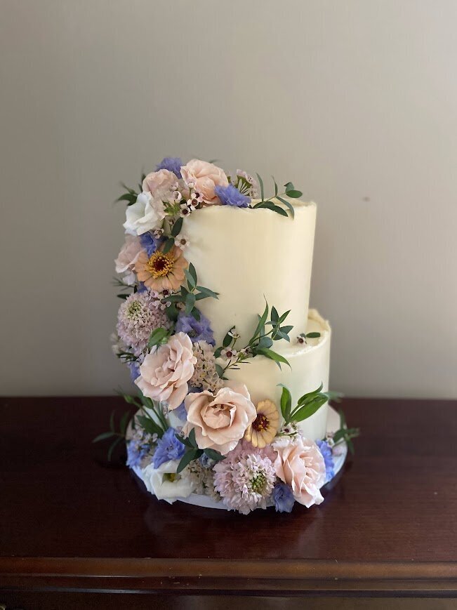 A two tier white cake is decorated with a cascade of fresh flowers in spring colors. 