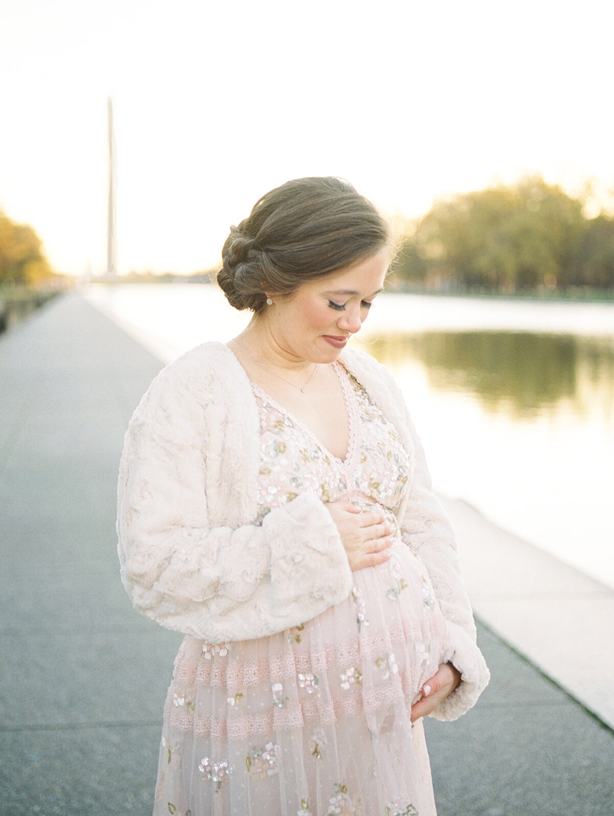 Pregnant mother with pink Needle & Thread dress and white fur shawl stands by the Reflecting Pool in DC.