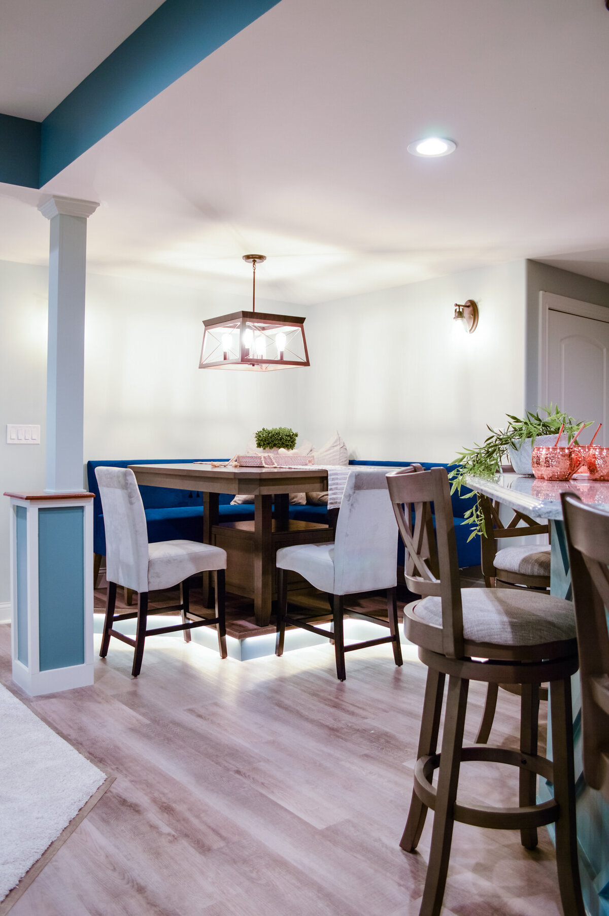 A view of a dining nook and kitchen island area in a well-lit space. The dining nook features a dark wooden table surrounded by a built-in blue banquette and two light gray upholstered chairs with dark legs. Under-cabinet lighting illuminates the banquette seating area. Above the table, a modern pendant light with a dark frame and clear glass panels hangs from the ceiling. To the right, a kitchen island with a light blue base and a patterned countertop is partially visible, with two Barstools with light gray fabric seats and cross-backs positioned around it. The floor is covered with light-colored wood-look planks. The walls are light gray, with a teal accent band near the ceiling. A white door is visible in the background on the right.
