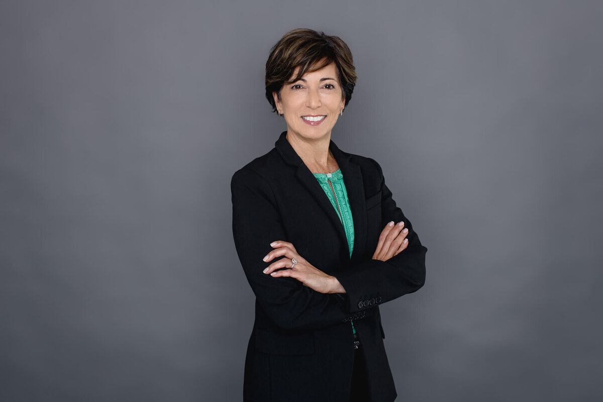 A woman with short, black hair facing the camera against a dark grey background, wearing a black blazer and crossing her arms at her chest, smiling.