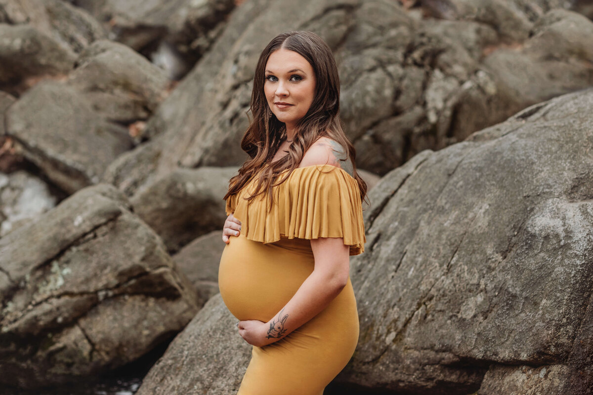 pregnant mother wearing a mustard maternity gown standing near large boulders near a stream