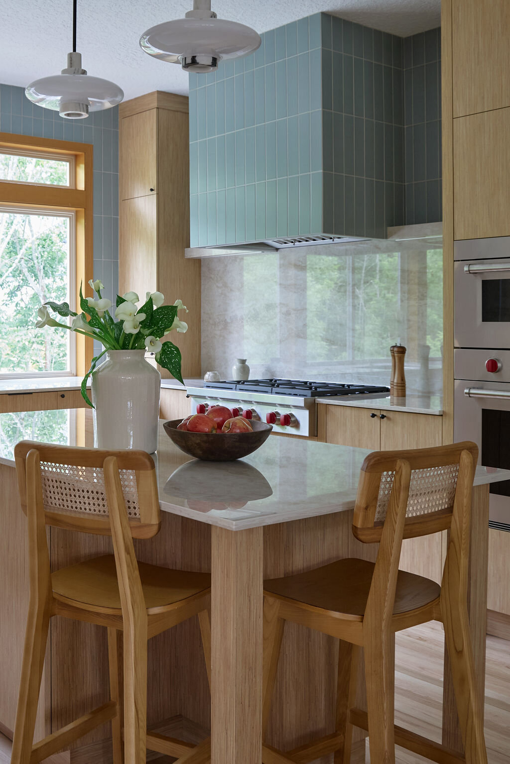 Kitchen with white oak cabinets, and slim simple black hardware. Taj mahal quartzite countertops that run up the backsplash to a quartz shelf with cookbooks and plants on it. Green subway tile backsplash above the shelf to the ceiling. Two sconces above the shelf that are modern, white and gold.