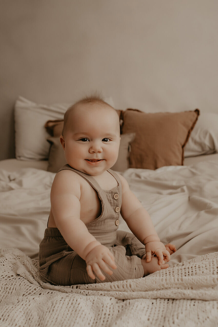 a 6 month old boy sits on a bed while smiling and looking at the camera