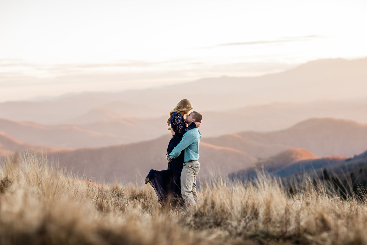 Young woman in a navy lace dress being lifted up by young man and embracing with panoramic view of Roan Mountain by Danielle Defayette Photography