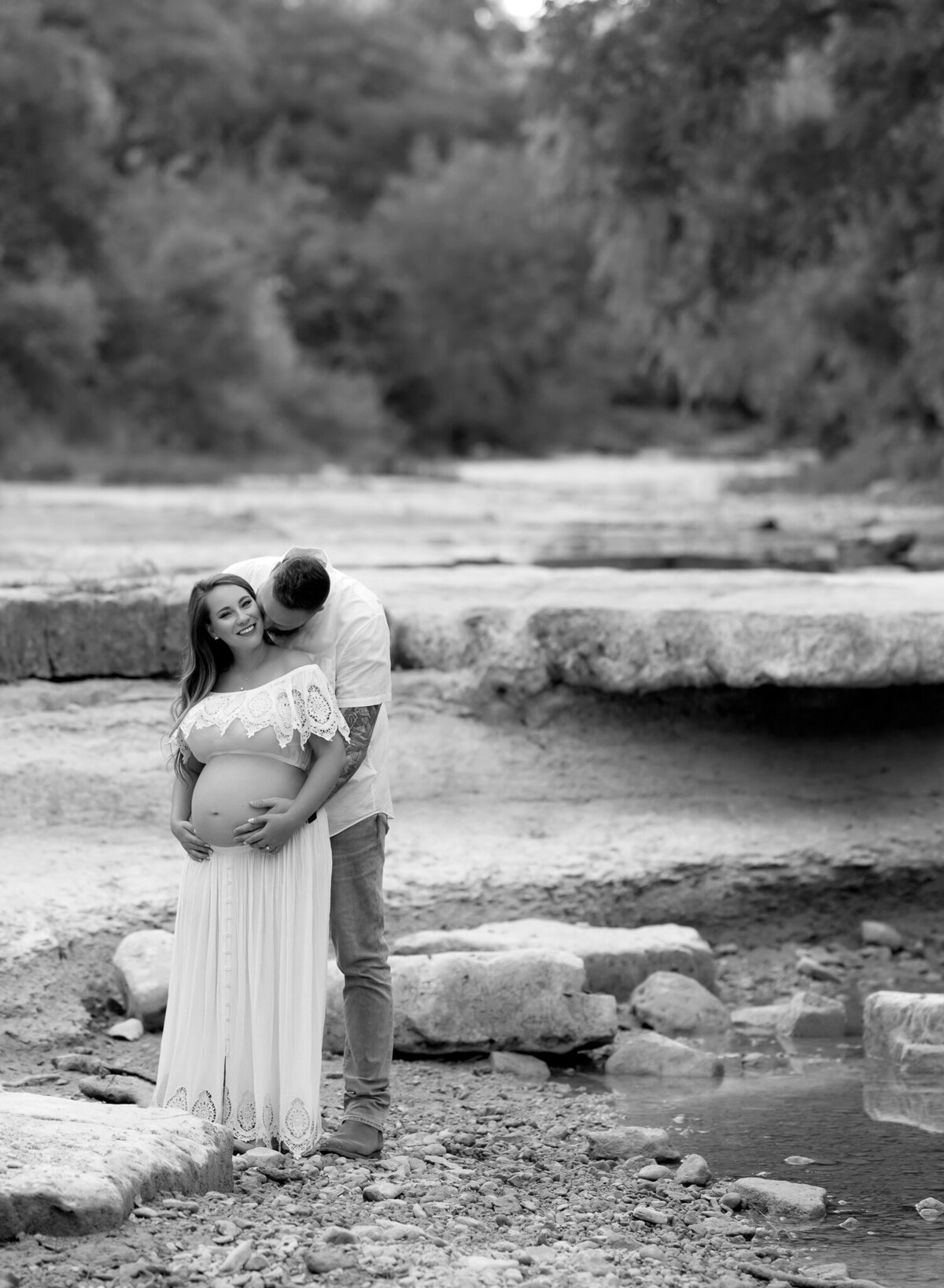Sweet maternity moment in Leander, Texas as a couple embraces near a rocky creek, with the mother-to-be in a white boho dress.