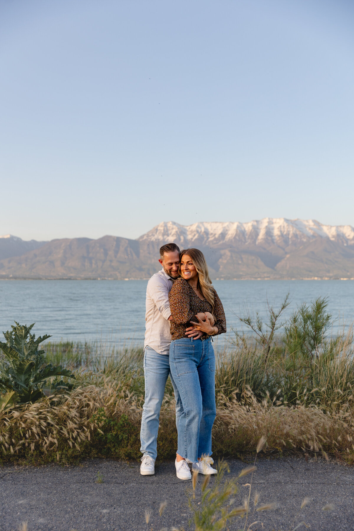 A man embraces his fiancée from behind, both smiling as they stand near Utah Lake. The serene blue water and distant mountains add to the picturesque setting.