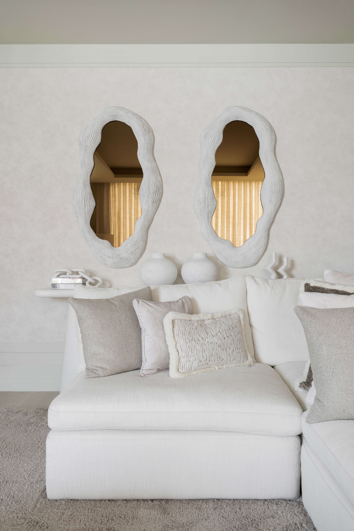 A spacious, modern living area with a focus on light, neutral tones. Behind the white couch is a white console table adorned with neutral colored decor. Above the table are two large, contemporary mirrors.