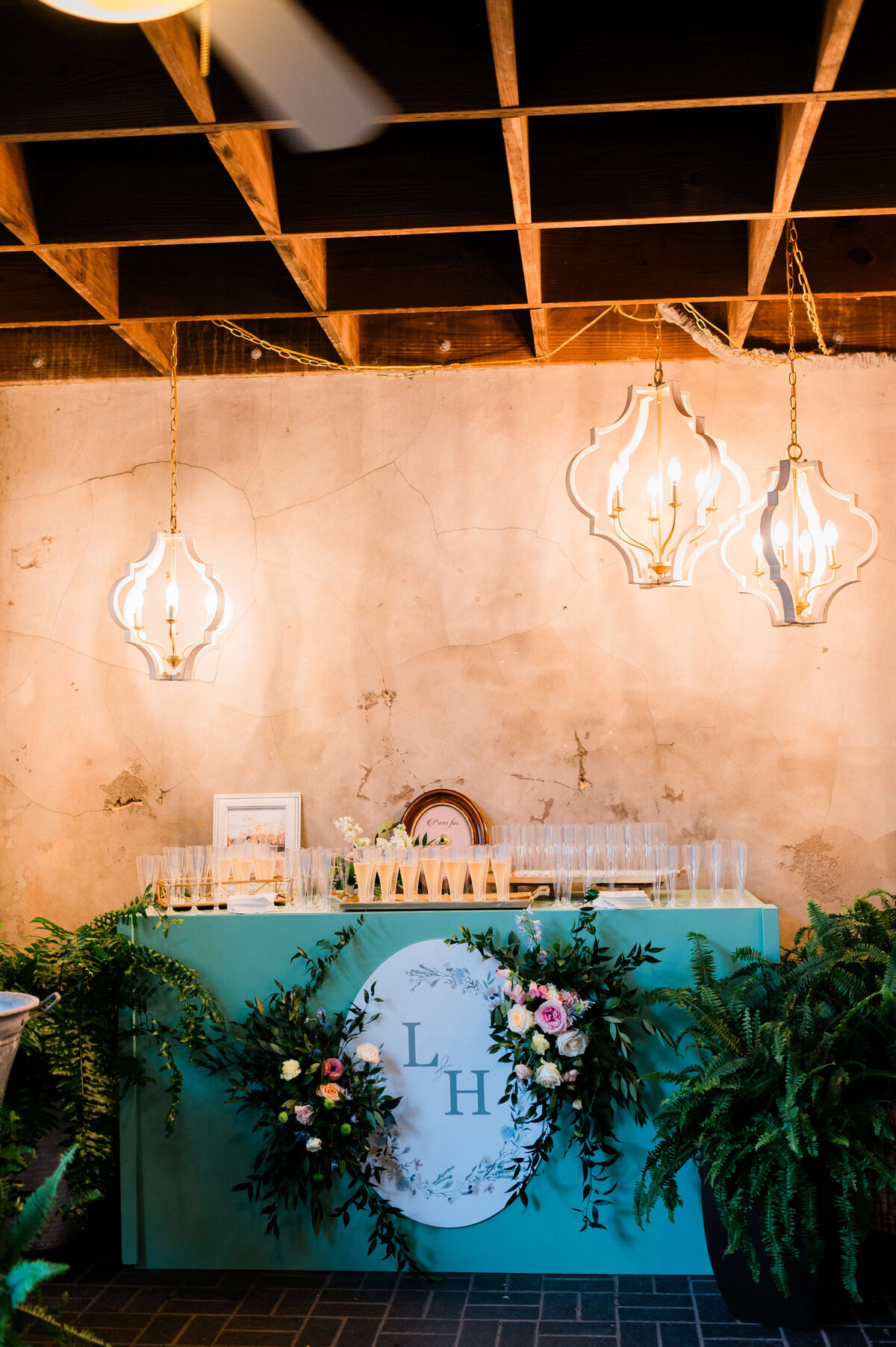 A green table decorated with a floral arrangement and ferns holds numerous filled champagne flutes. Behind the glasses is a round clock against a textured, beige wall.