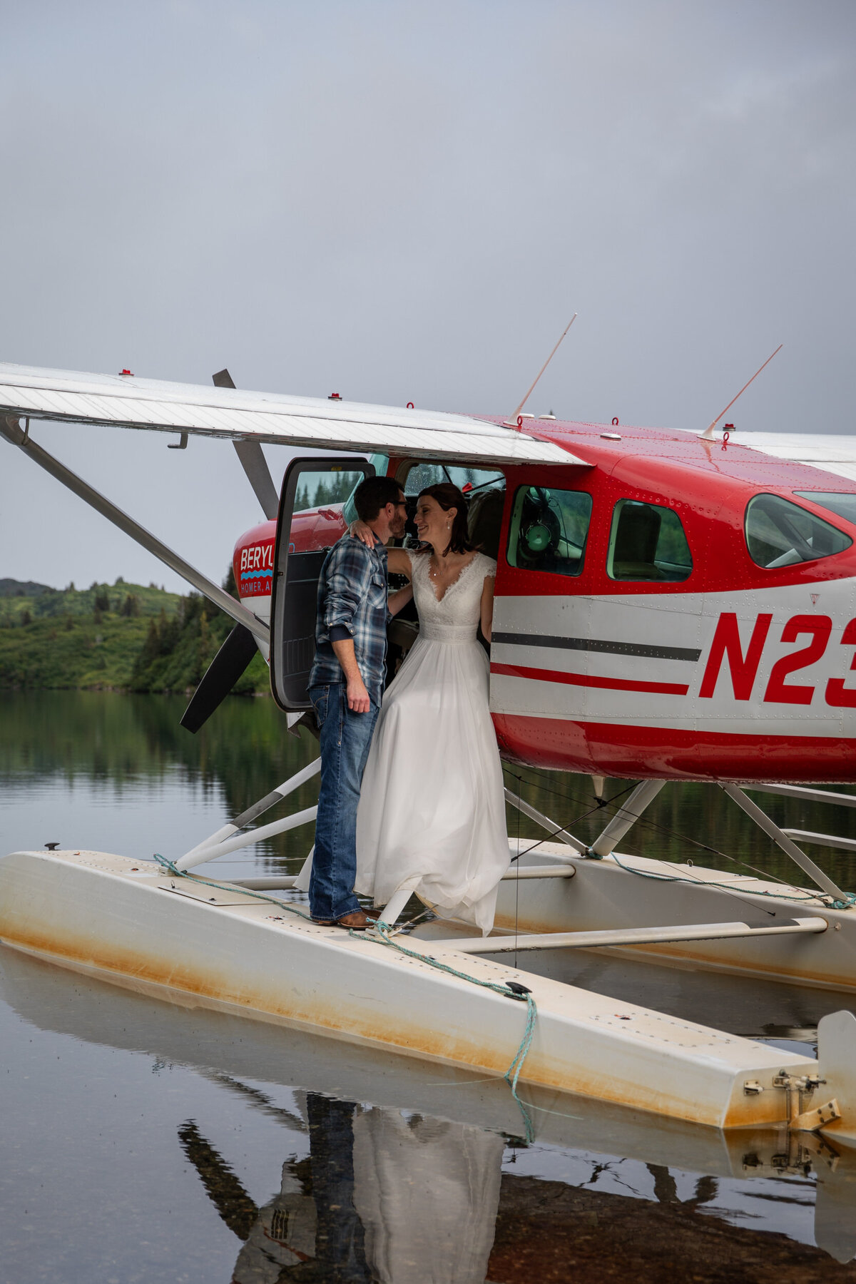 A bride sits in the open door of a float plane while her groom stands in front of her.