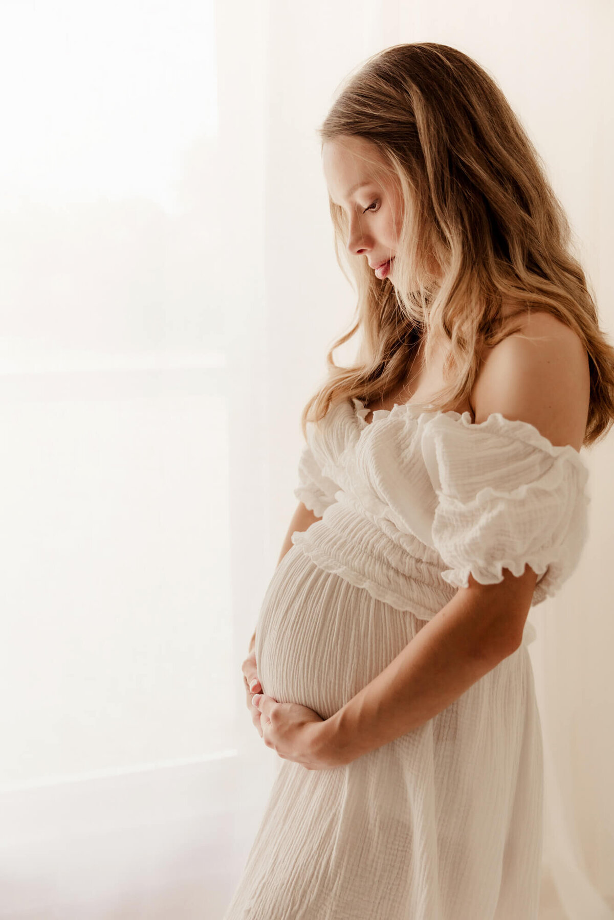 Woman holding her baby bump while looking down and wearing a white off the shoulder ruffled dress, OKC.