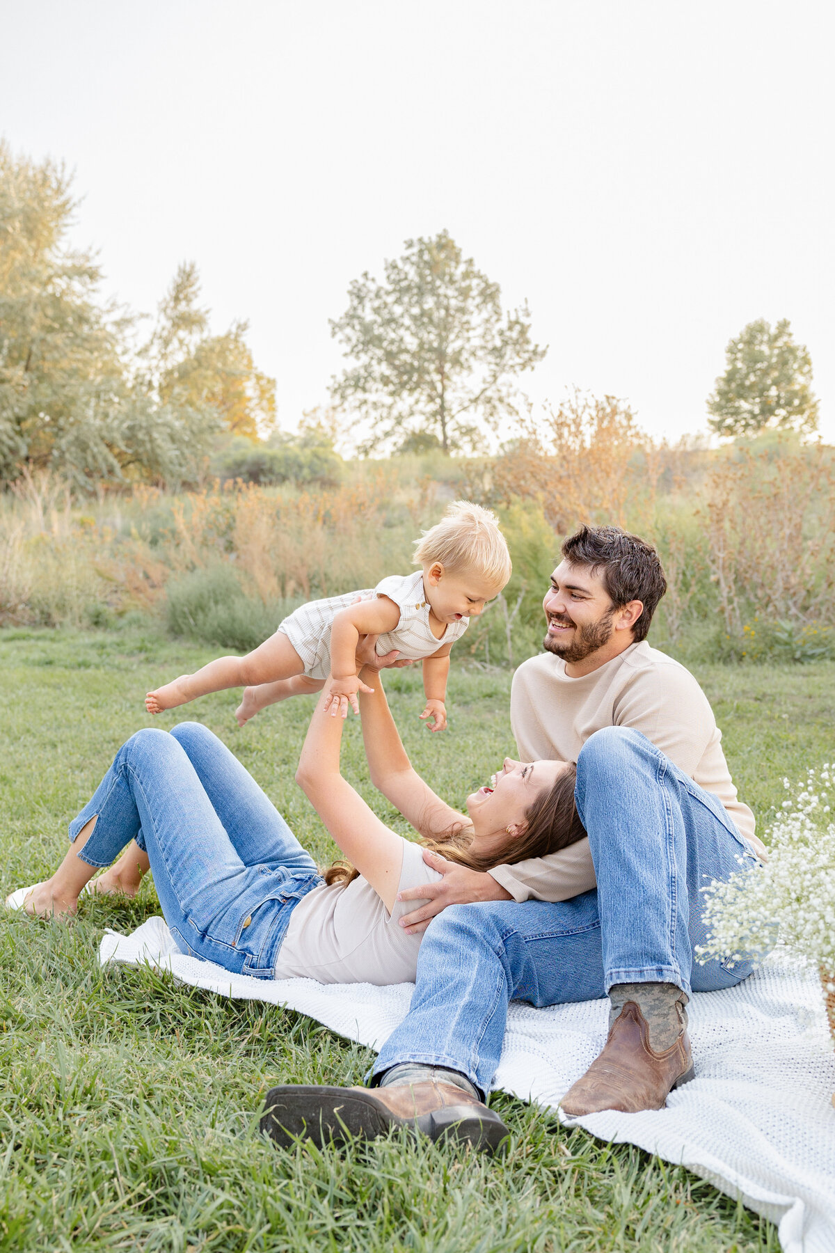 A mother lies on a blanket in a grassy field while holding her baby in the air as the father watches with a smile. A joyful family moment captured by Cassie Lee Photography in Eagle Mountain, Utah.