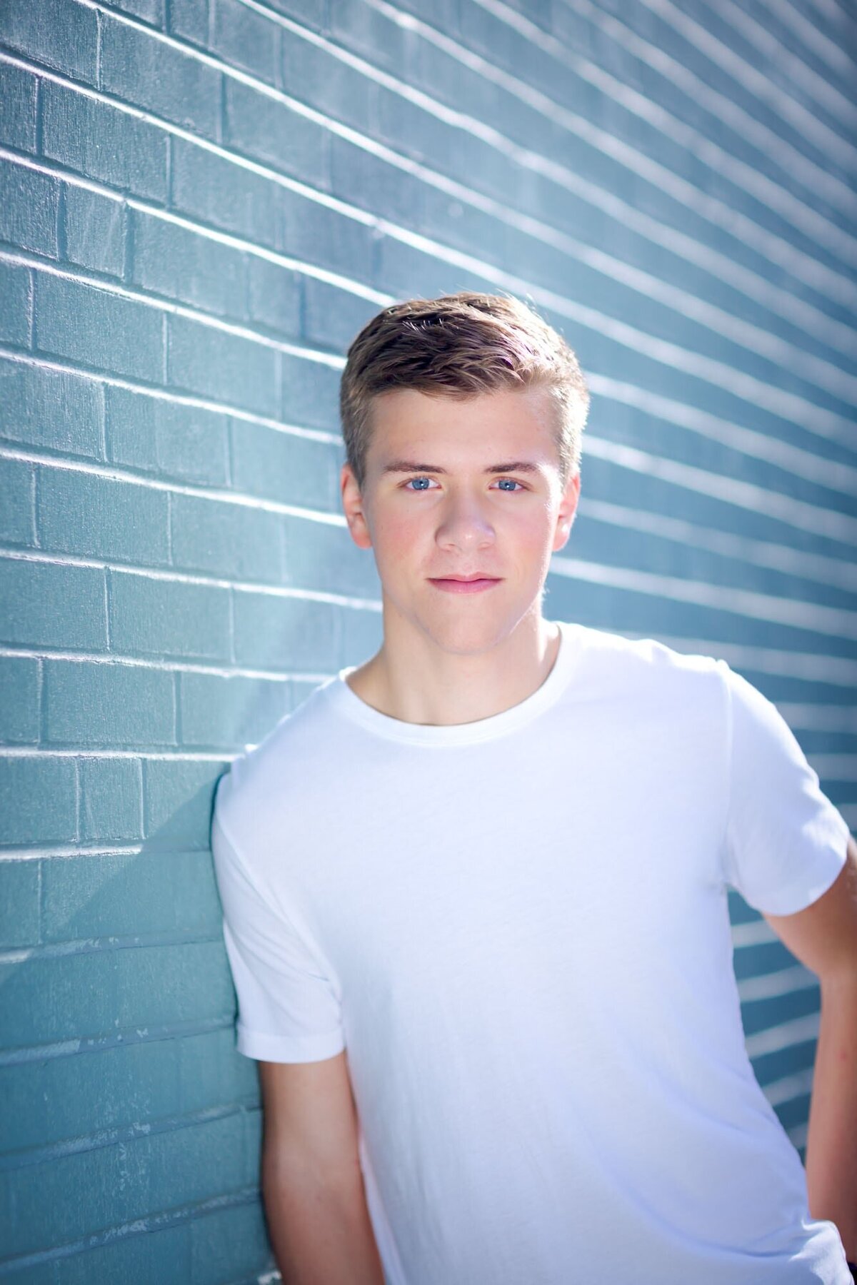 Senior guy in a white shirt leaning against a blue brick wall.