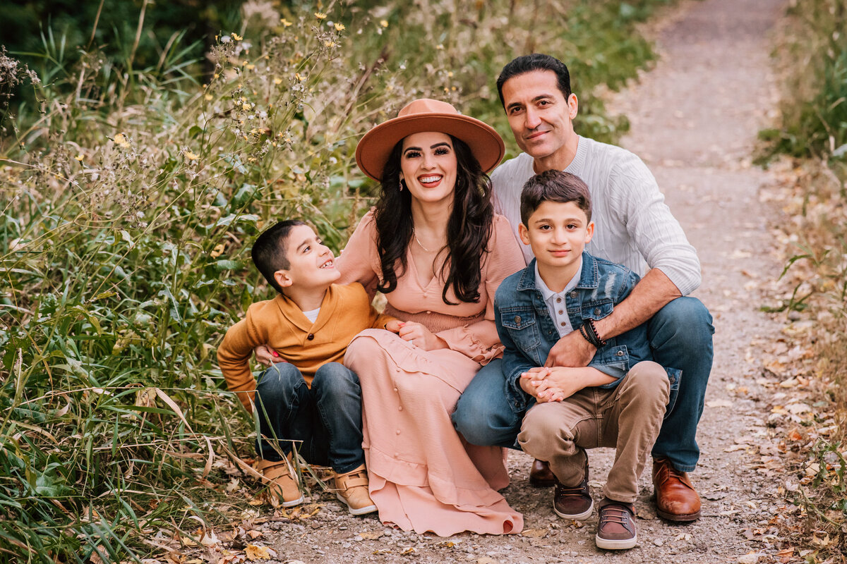 A family of four sits together on a trail surrounded by tall grass and wildflowers. The mother is wearing a hat and a long dress, while the father and two sons sit close, all smiling warmly at the camera.