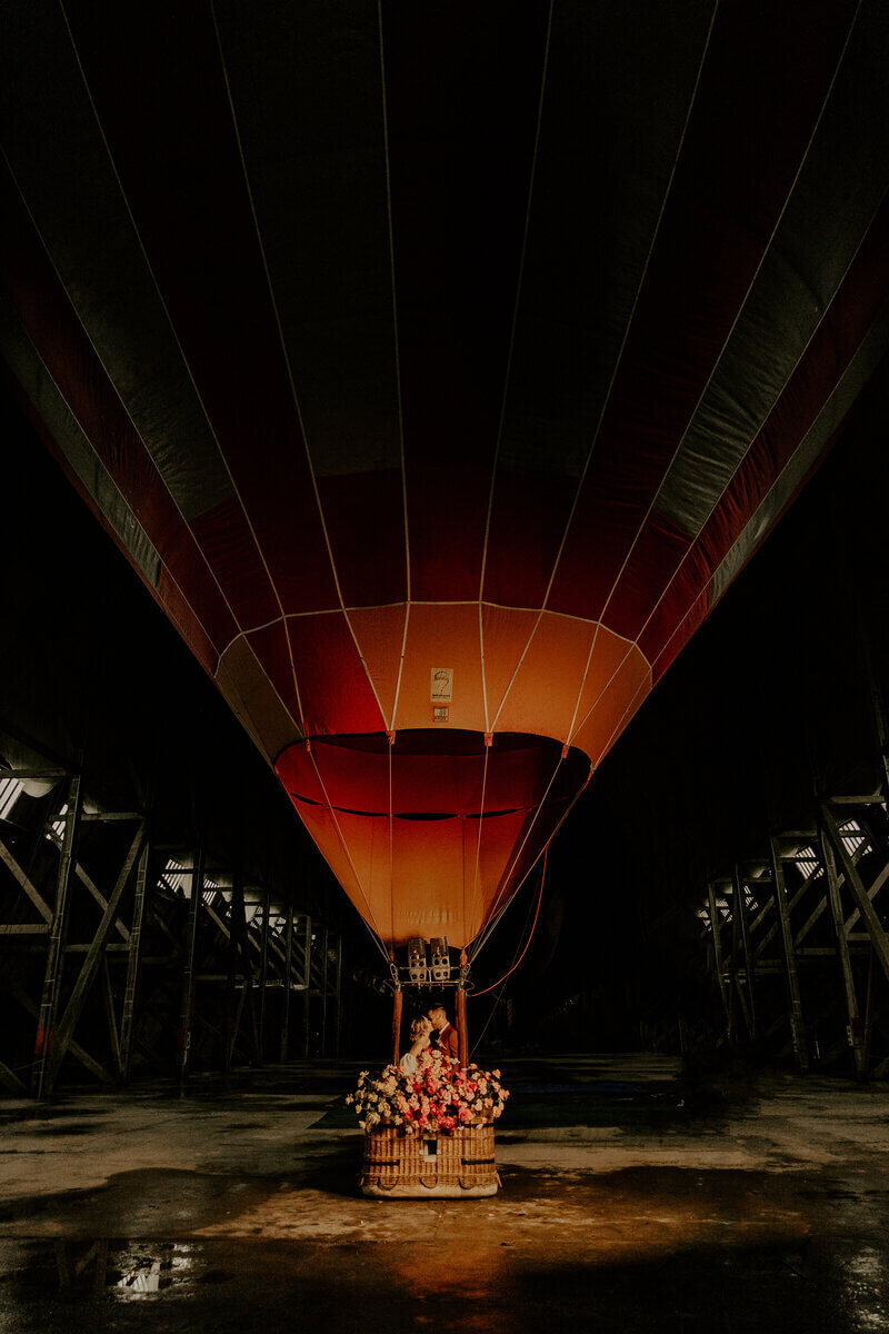 Mariés au loin échangeant un bisou dans une montgolfière au ballon rouge garnie de fleurs lors d'un shooting photo mariage.