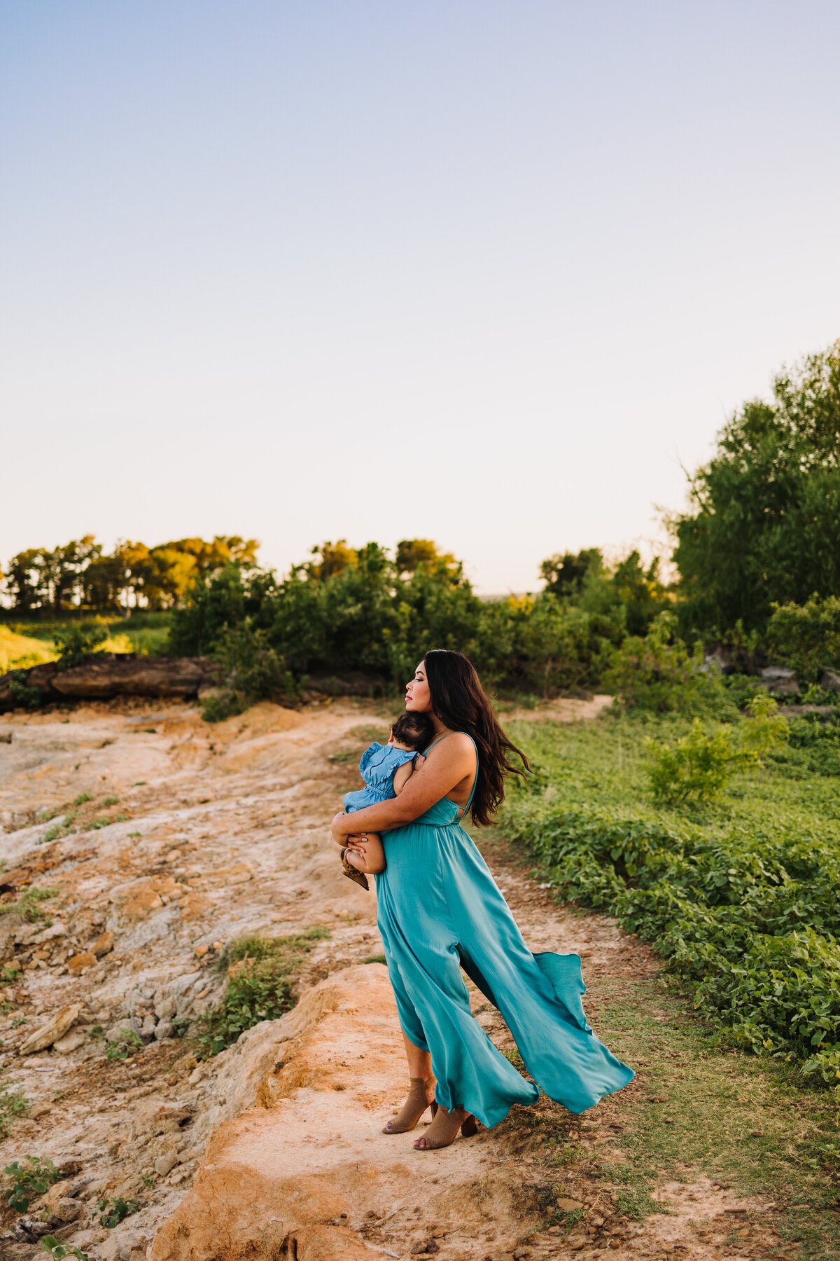 Photo of a woman in an aquamarine dress carrying a baby in her arms. She is in profile and with her eyes closed, she is standing on some rocks and behind her there are some green trees