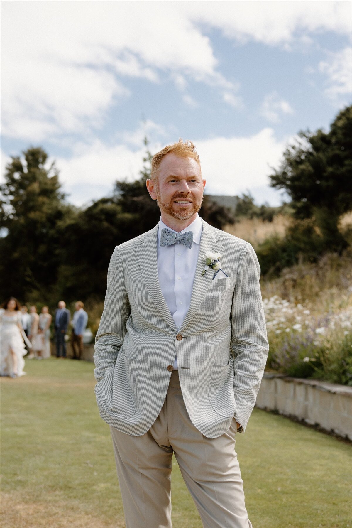 Groom in a pale seersucker jacket and floral boutonnière smiling with hands in pockets before the ceremony at a Lake Hāwea wedding.
