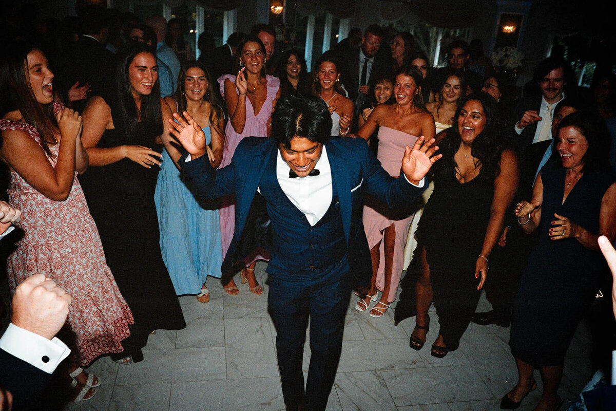 A man in a navy suit and bow tie dances energetically in the center of a lively group of smiling, well-dressed people at a formal indoor event, as an NJ wedding photographer captures the joyful moment while the group cheers him on.