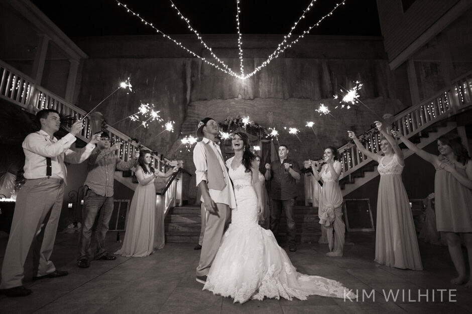 A bride and groom smile and walk hand in hand through a courtyard at at night, surrounded by guests holding lit sparklers and celebrating their wedding.