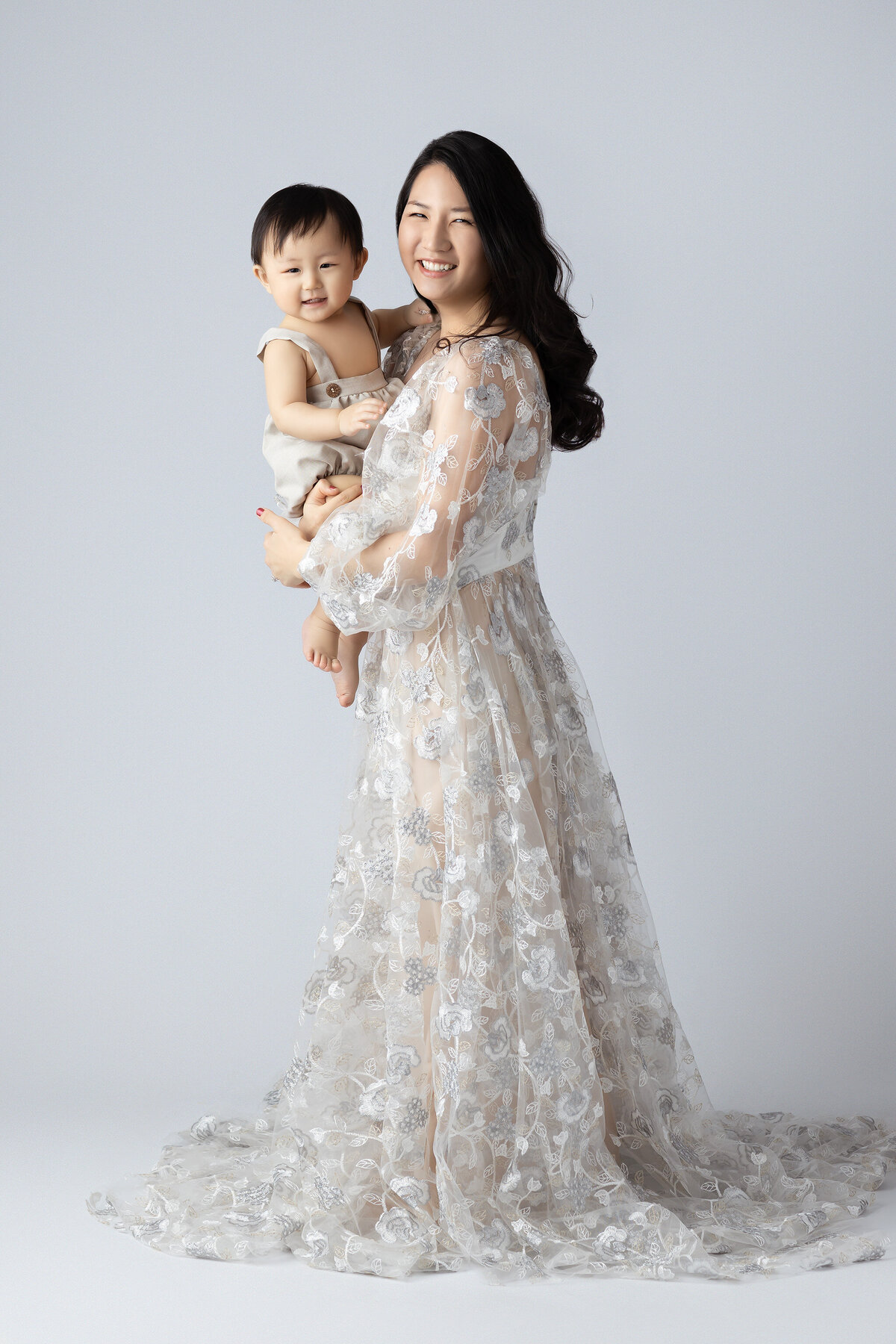 Dallas baby photographer captures a mother smiling with her son in a soft, flowing floral gown and neutral background.