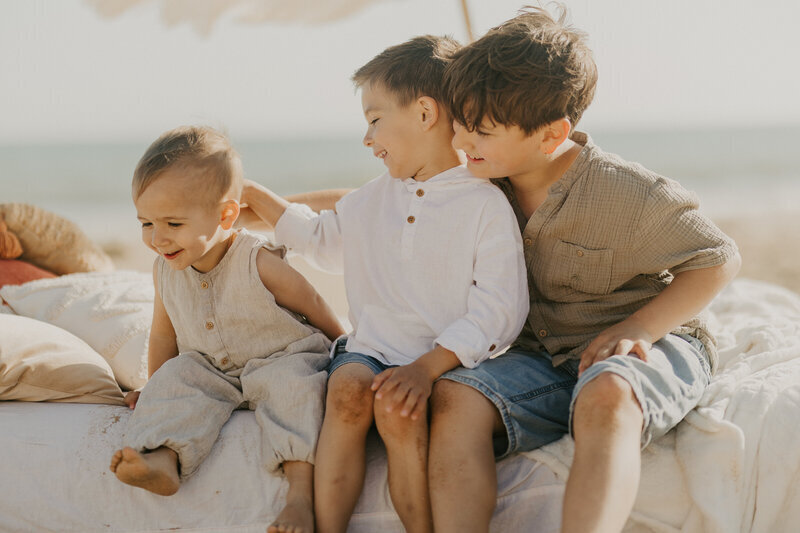 Trois petits garçons d'âges différents, complices, sur un décor cosy de matelas et coussins à la plage lors d'un shooting photo famille en Vendée.