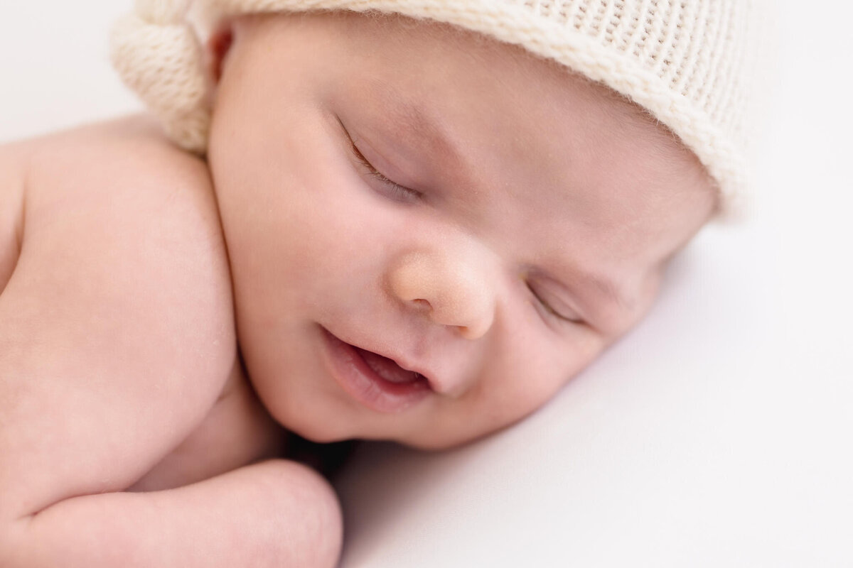 A close-up of a sleeping baby wearing a knitted cap. The baby is smiling gently, with eyes closed and head resting on its arm.