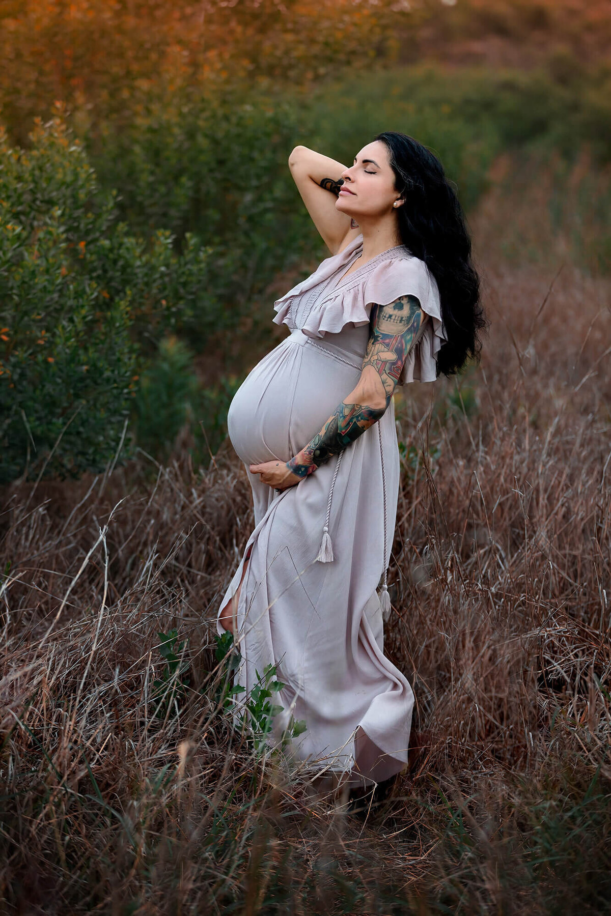 Beautiful mom to be looking up while holding her belly with tattoos in her arm on a field in Pearland, Texas.