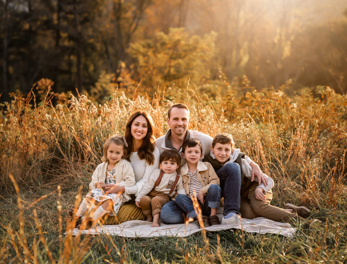 A mom, dad and their four children snuggle on a blanket in the yellow fall grass