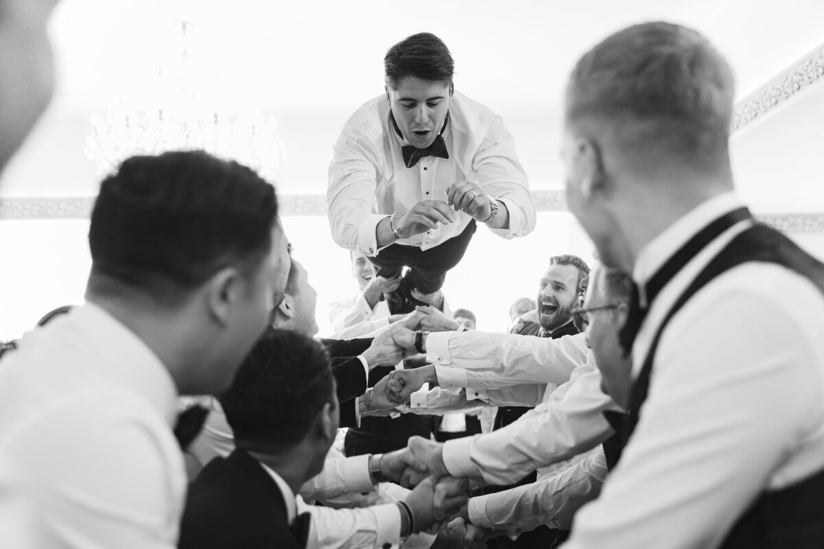 A group of men dressed in formal attire lift a groom in the air during a celebration at Froyle Park.  The groom is smiling and wearing a bow tie and white shirt. The scene is joyful and energetic, captured in black and white.