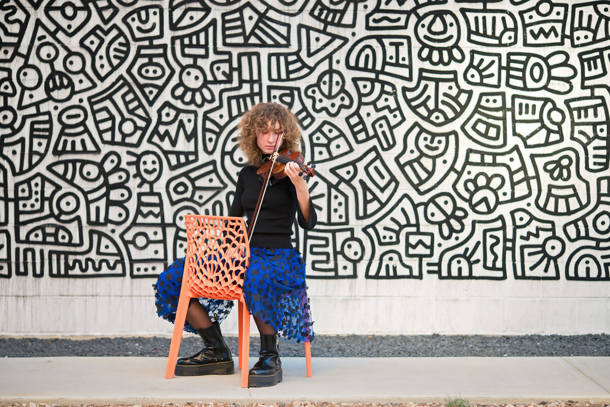 A high school senior girl plays her violin in front of an urban mural.