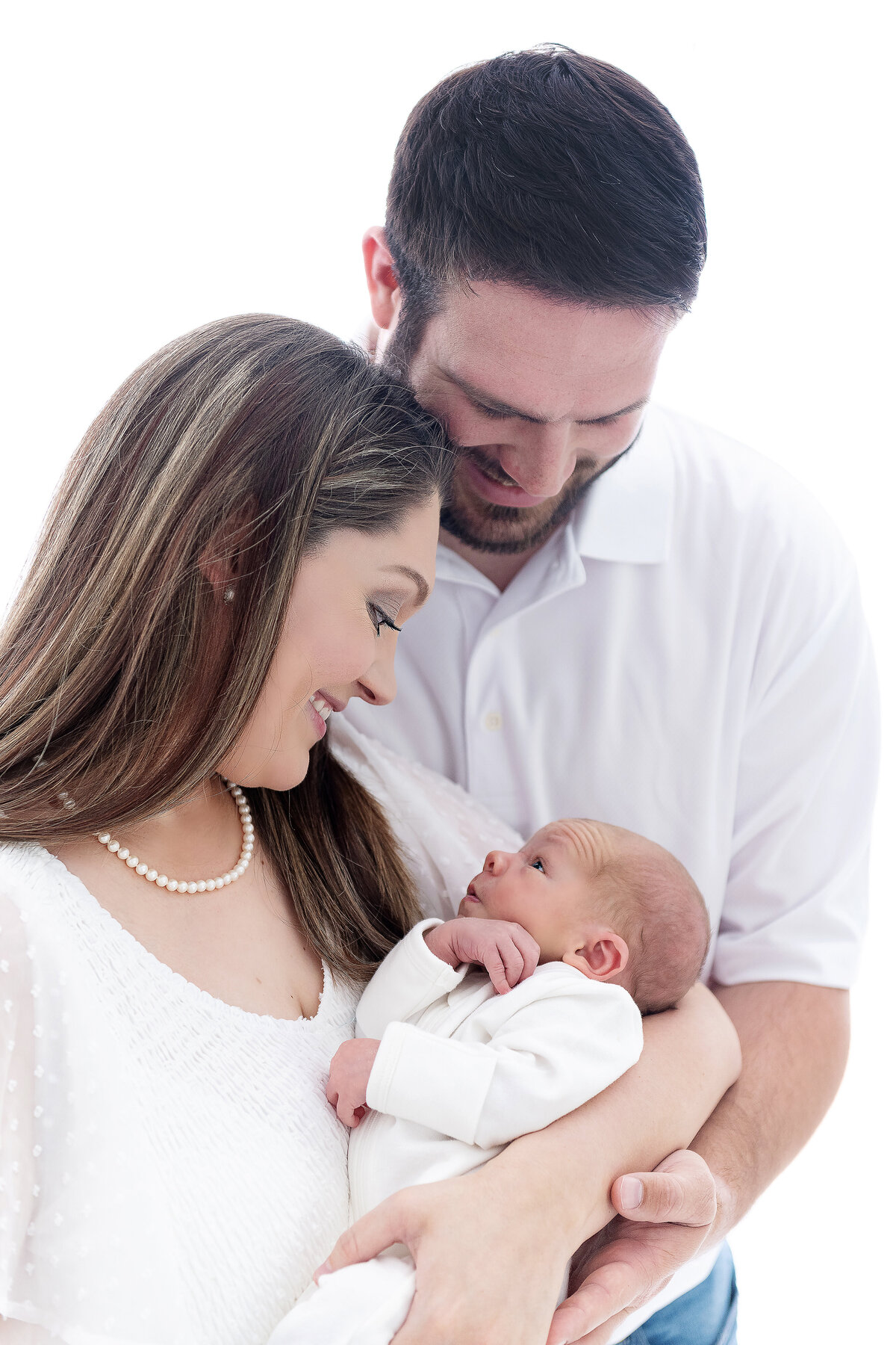 Happy parents smile down at their awake newborn baby while they cradle it standing in a studio