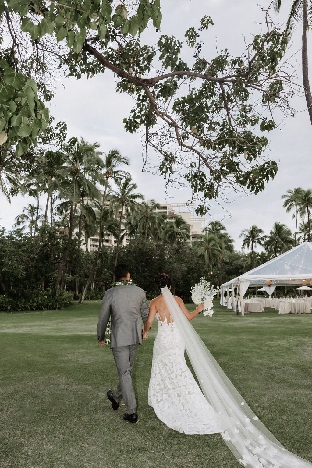 bride and groom walking, bride has a long train and there is a wedding tent in the background