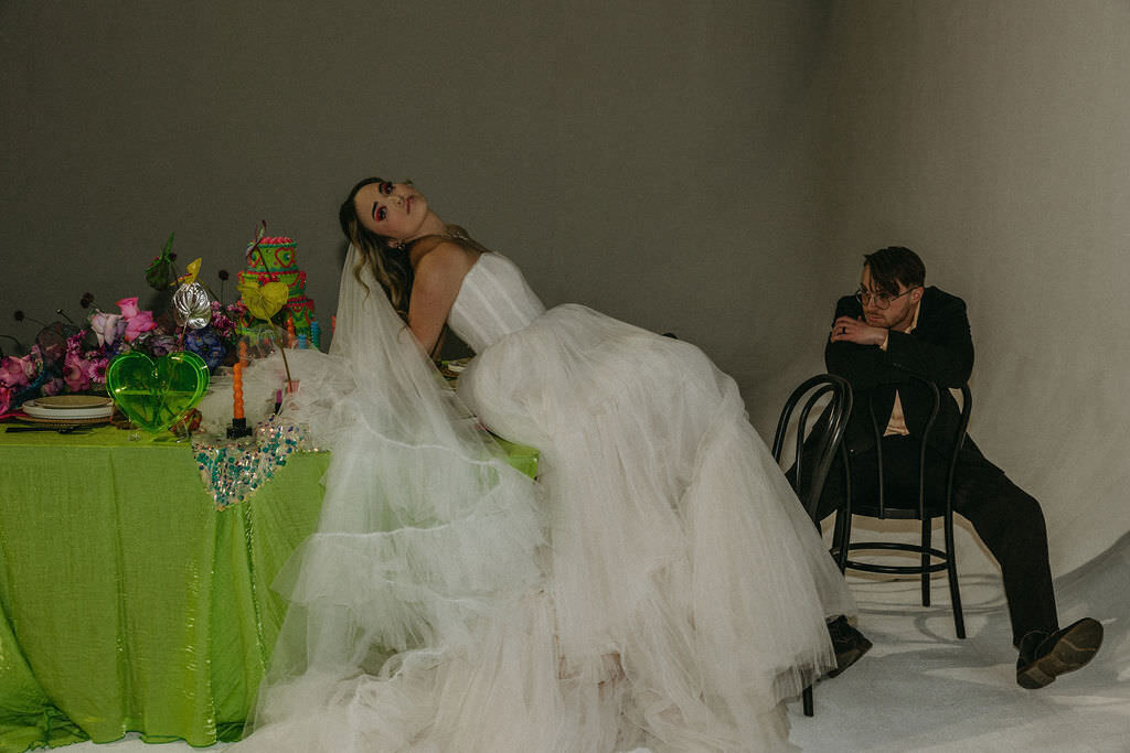 A person in a wedding dress sitting on a table with their partner in a chair next to them.