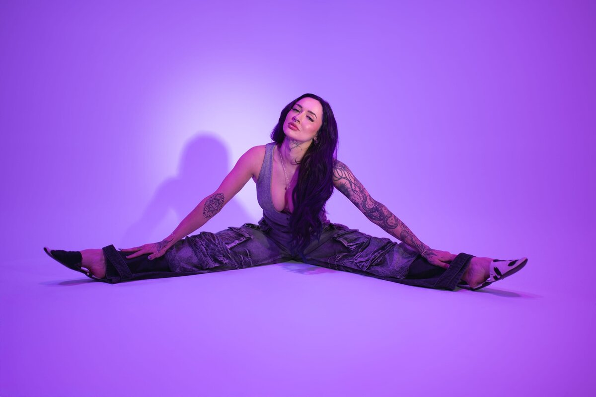 Woman sits in a straddle position with her head cocked to one side and looks at the camera in front of a purple backdrop.