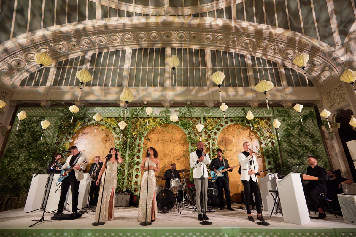 The photo was taken at The New York Public Library in New York City by photographer Shawn Connell. The event was captured by Aaron Navak Films, who provided videography services. A live band was performing on a stage adorned with green foliage and yellow hanging lanterns, set against a backdrop of large golden panels. The performers included singers and musicians with various instruments, all under intricate ceiling architecture.