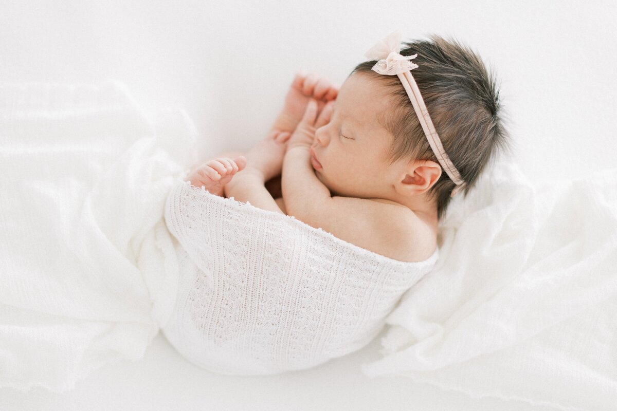 Newborn baby girl with pink bow and a lot of brown hair curled up with white swaddle sleeping