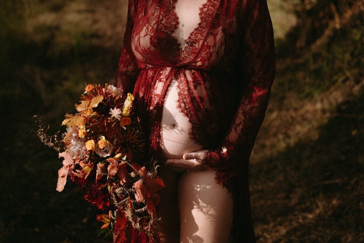 Femme enceinte en robe de dentelle rouge, une main sur son ventre rond apparent et l'autre tenant un bouquet automnal. Photo par Kamescop.