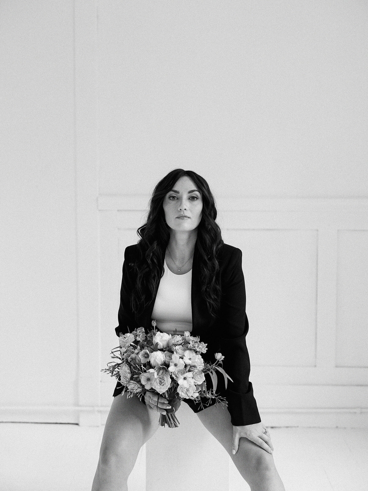 A woman in a black blazer and white top sits on a stool in a minimalist room, holding a bouquet of flowers, embodying the elegance one would expect from a wedding planner logo.