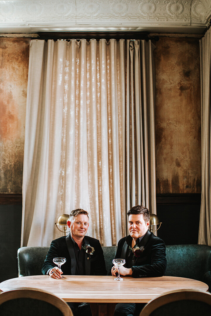 Two grooms wearing black tuxedos hold drinking glasses while sitting on a green leather couch.