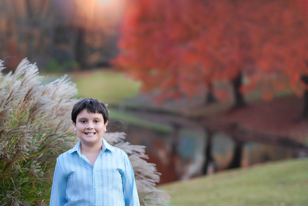 Little boy standing in a park for portrait. Is is looking and smiling at the camera. In the background, the fall trees are a vibrant red and the colour is reflecting off of a pond. Captured by Liza Nicole Photography, Philadelphia and Main Line family photographer.