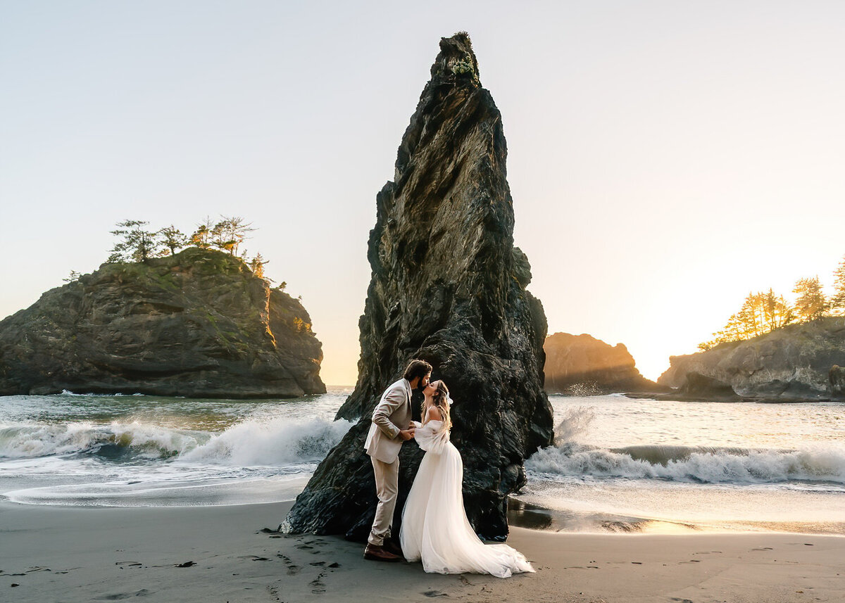 During their Oregon coast elopement, a couple kisses in front of a jagged sea -stack as a vivid sunset paints the sky behind them and waves roll onto the beach