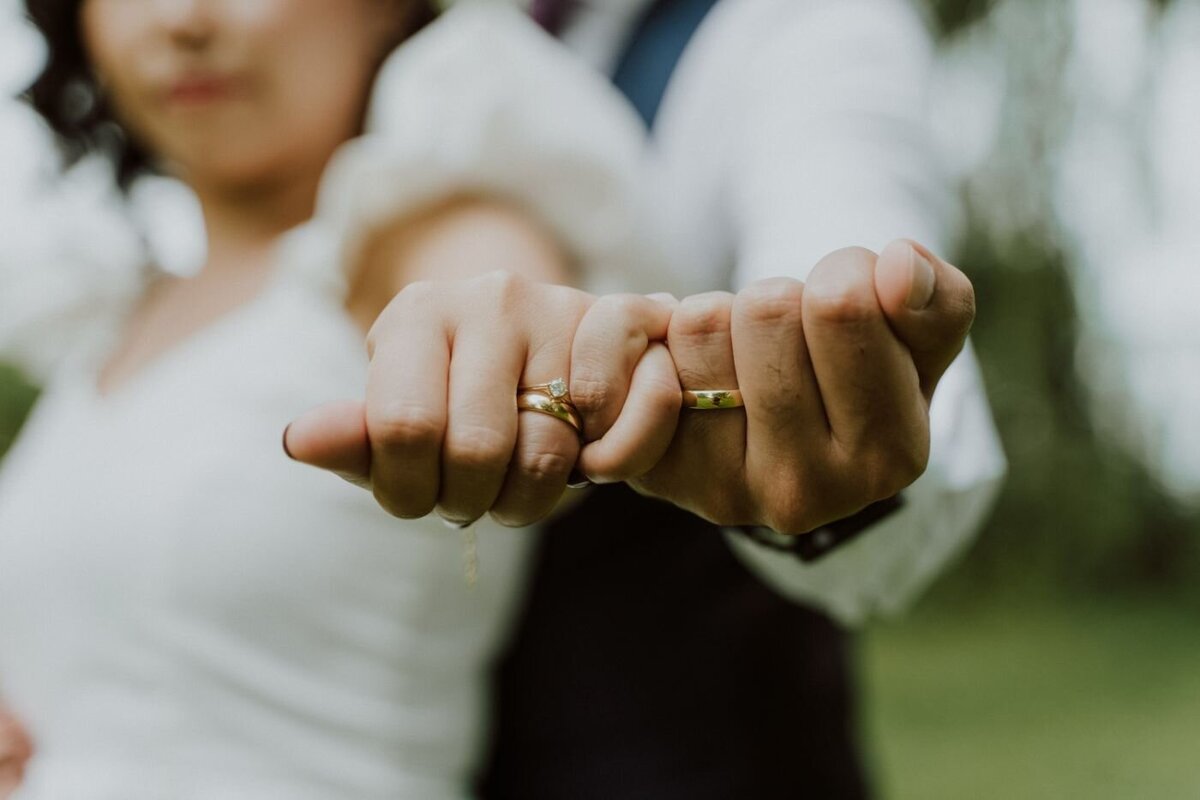  A close-up shot focusing on the hands of a newly engaged couple. They are holding hands, and their engagement and wedding rings are clearly visible on their left hands. The woman's hand is on top, showcasing a golden ring with a large diamond. The man's hand is underneath, with a simple gold band. The background is a soft, out-of-focus green, suggesting a park or field.