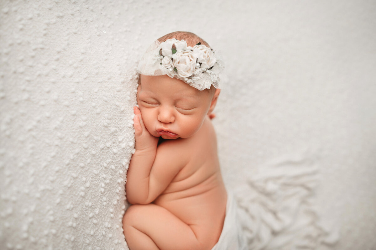 newborn baby girl posing on her belly wearing white headband on white backdrop
